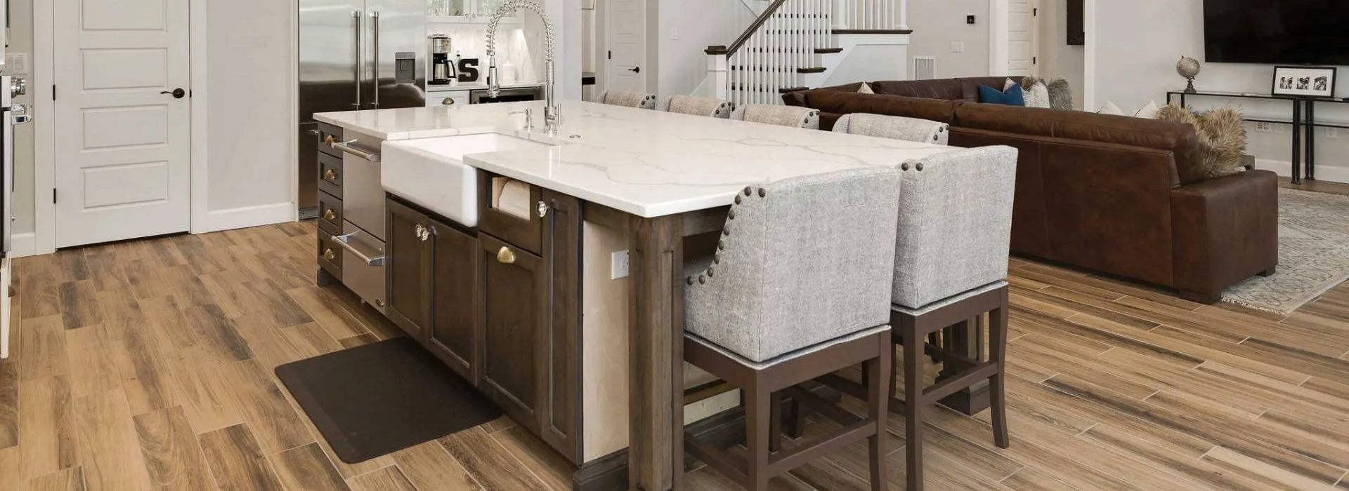 Open-concept kitchen with white island, gray chairs, hardwood floors, and brown sofa in the background
