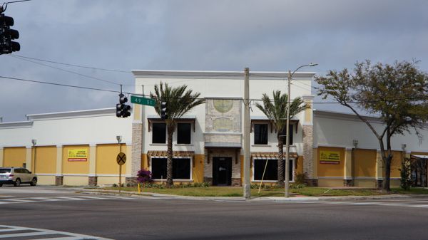 White commercial building with palm trees at a street corner under a cloudy sky
