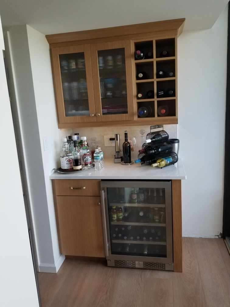 Wooden home bar cabinet with glassware, bottles, and a wine fridge against a white wall