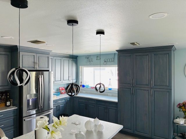 Blue kitchen with dark cabinets, stainless fridge, island, and white dining table under pendant lights.