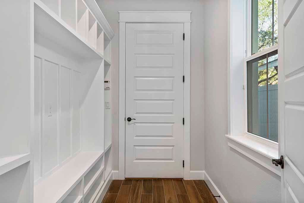 Bright white hallway with paneled door, built-in cabinets, and window above a hardwood floor.