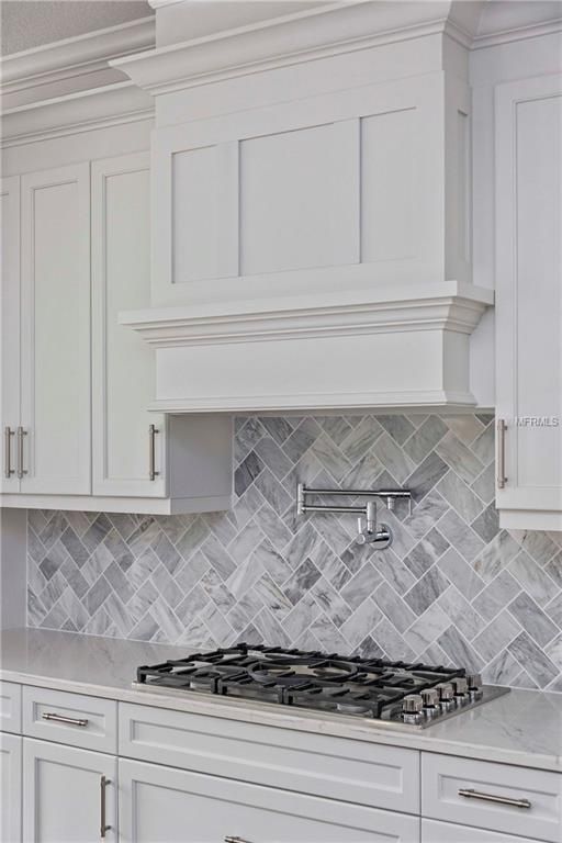 White kitchen with gray herringbone tile backsplash, gas cooktop, and range hood above the stove.