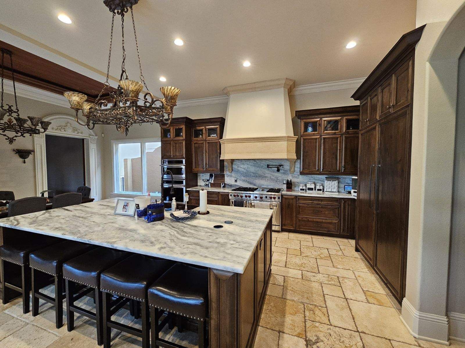 Modern kitchen with dark wood cabinets, large marble island, bar stools, and beige tile floor