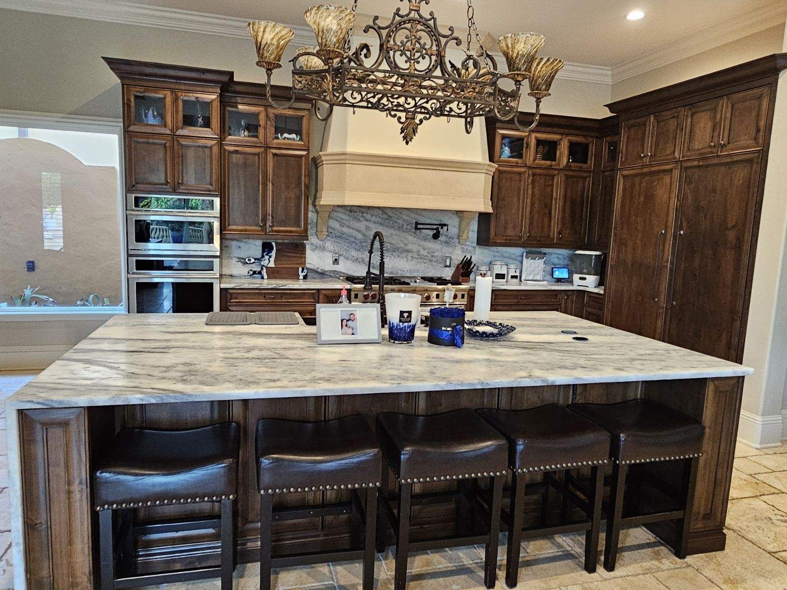 Luxury kitchen with dark wood cabinets, large marble island, and four bar stools under ornate chandelier