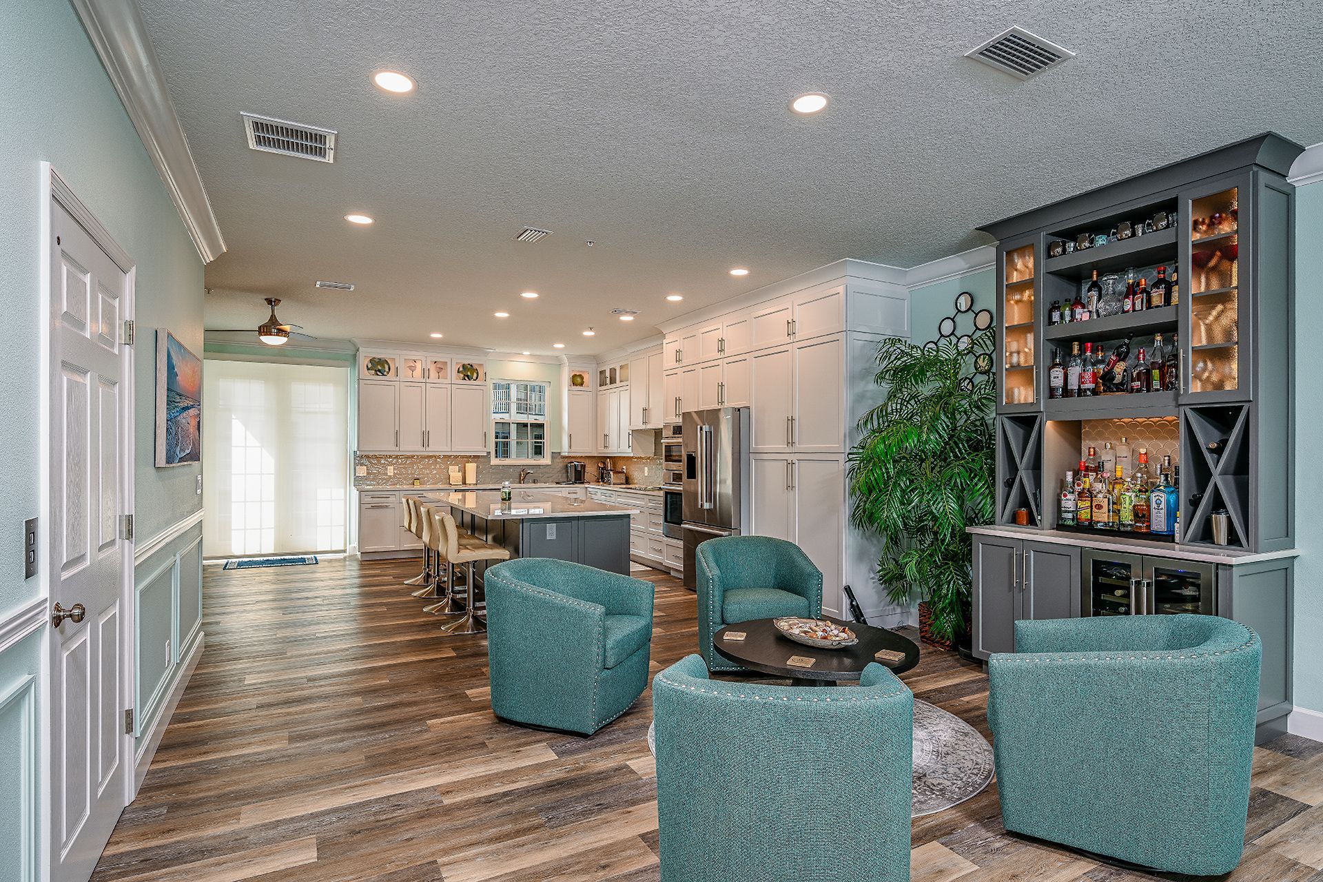 Bright lounge with teal chairs, wood floors, kitchen area, and shelves in a modern open space.