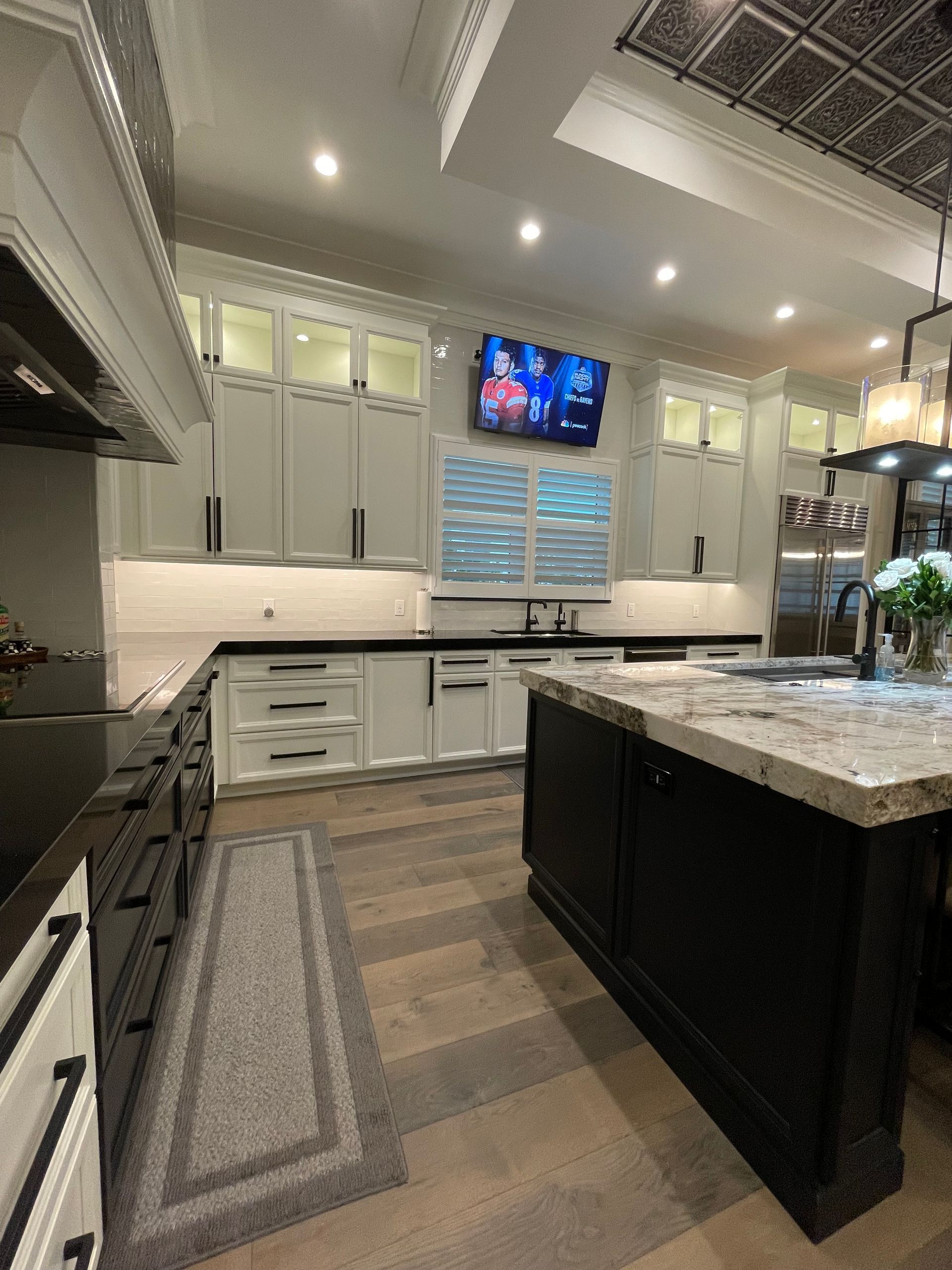 Bright modern kitchen with white cabinets, black island, granite counters, and a wall-mounted TV