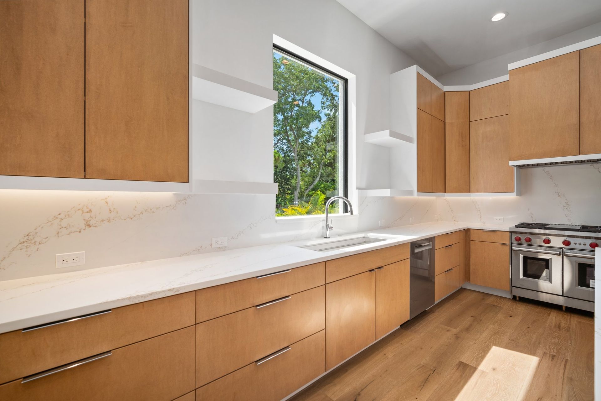 Modern kitchen with light wood cabinets, white countertops, stainless stove, and a window over the sink.