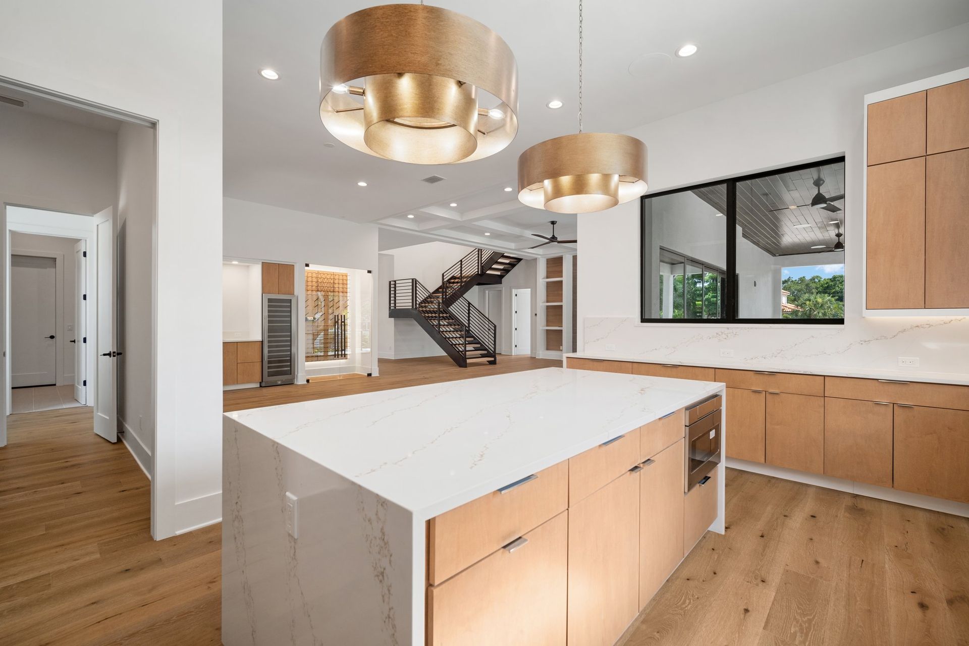 Modern kitchen with white island, wood cabinetry, gold pendant lights, and staircase in background
