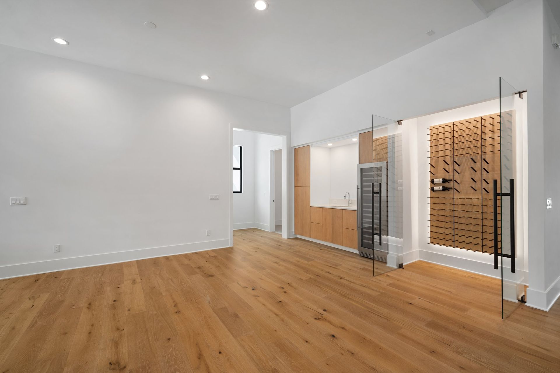 Empty modern room with light wood floors, white walls, and a glass wine display area