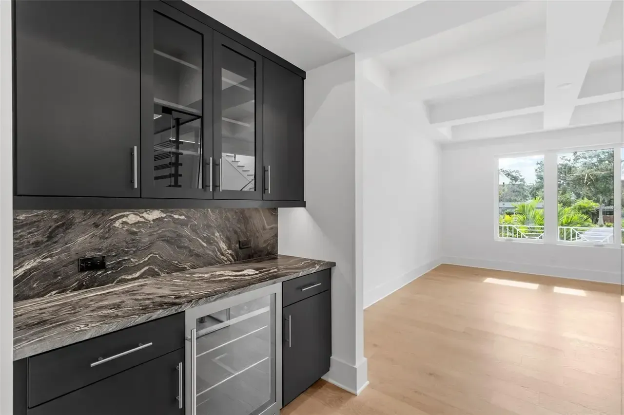 Modern kitchen with dark cabinets and marble backsplash opening to a bright empty room with wood floors
