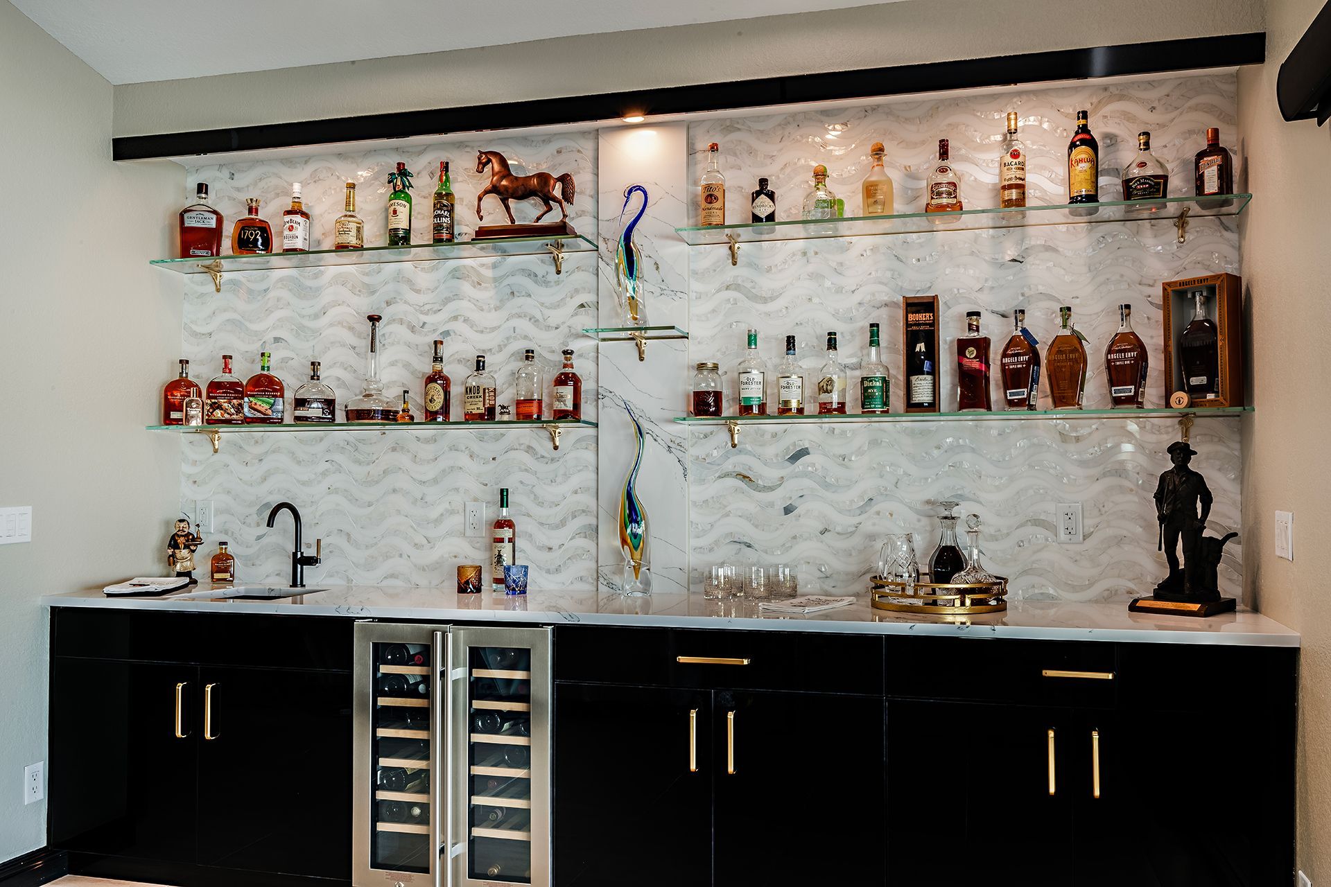Home bar with shelves of liquor bottles, glassware, sink, and dark cabinets under a marble backsplash.