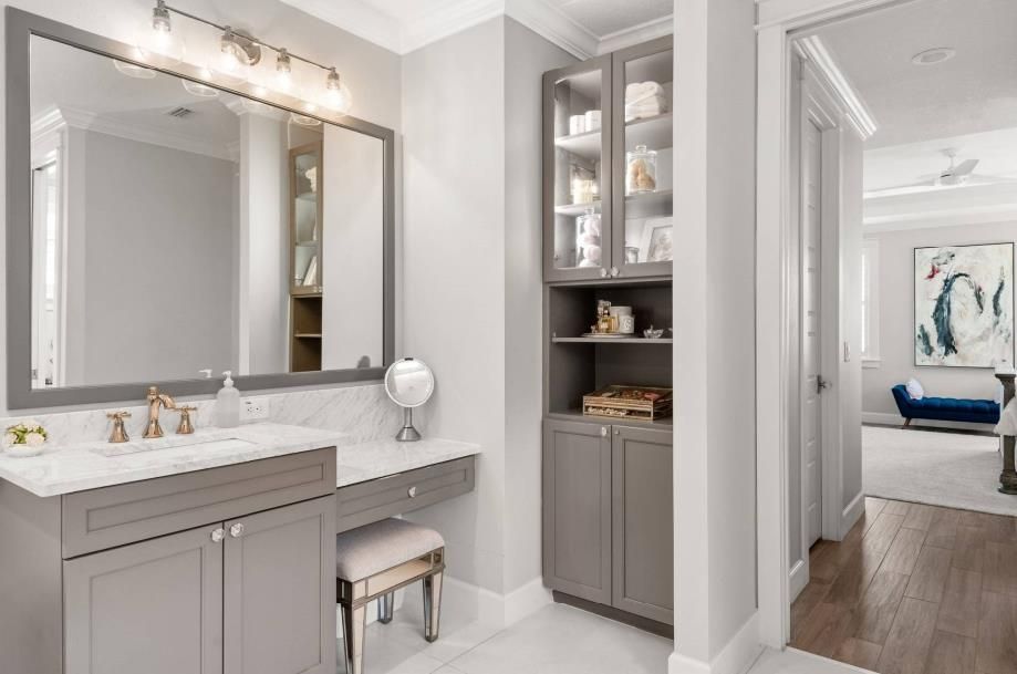 Bright gray bathroom vanity with large mirror, lighted wall sconces, and a built-in cabinet beside a doorway