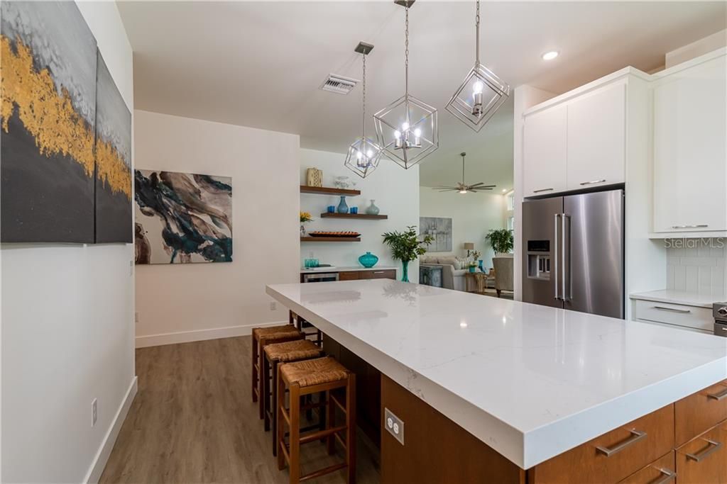 Bright modern kitchen with white island, pendant lights, wood stools, and stainless steel refrigerator.