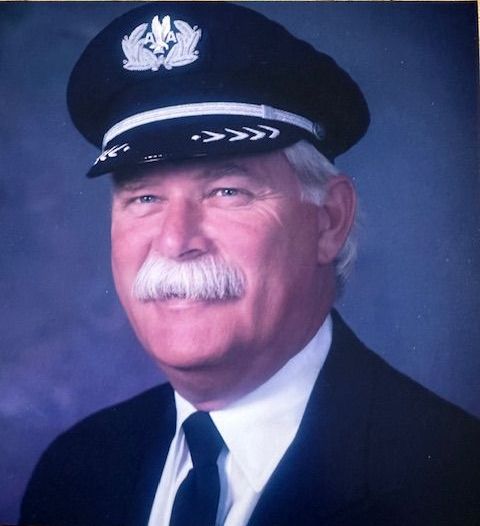 A man in a pilot's hat and uniform, smiling, with a white mustache, posing for a portrait.