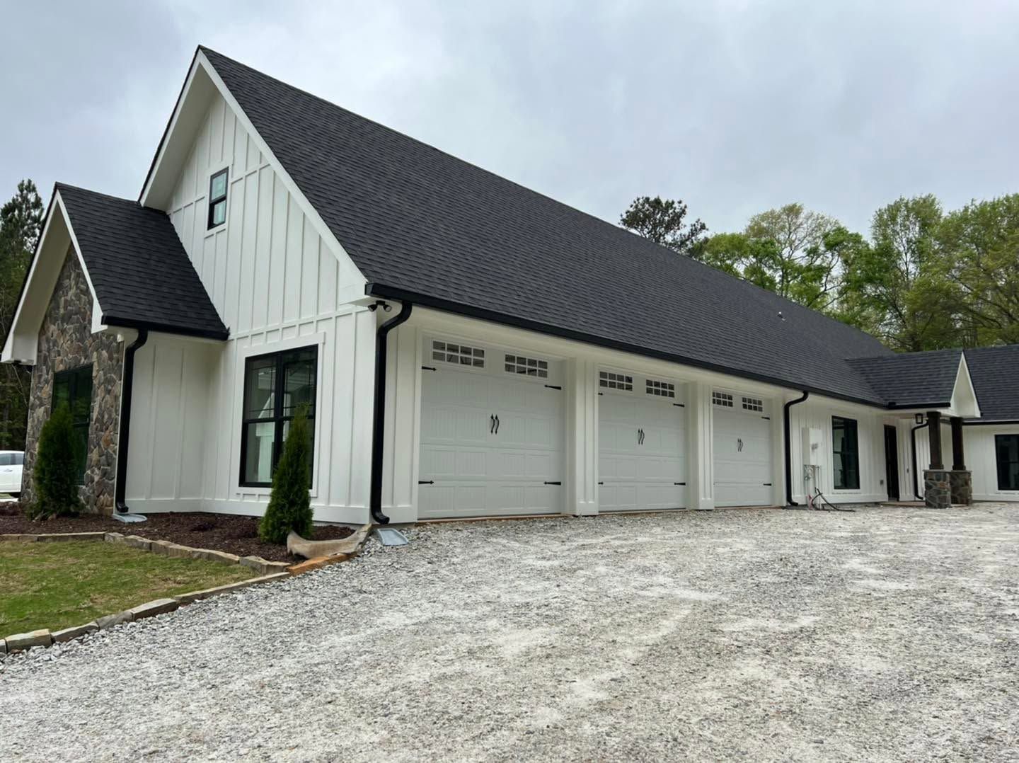 White house with black roof and three garage doors on a gravel driveway under cloudy skies
