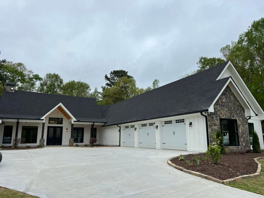Large white house with black roof, multiple garage doors, and a curved driveway under a cloudy sky
