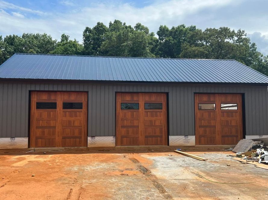 Three brown garage doors on a metal building with a gray roof, set on a dirt lot with trees behind.
