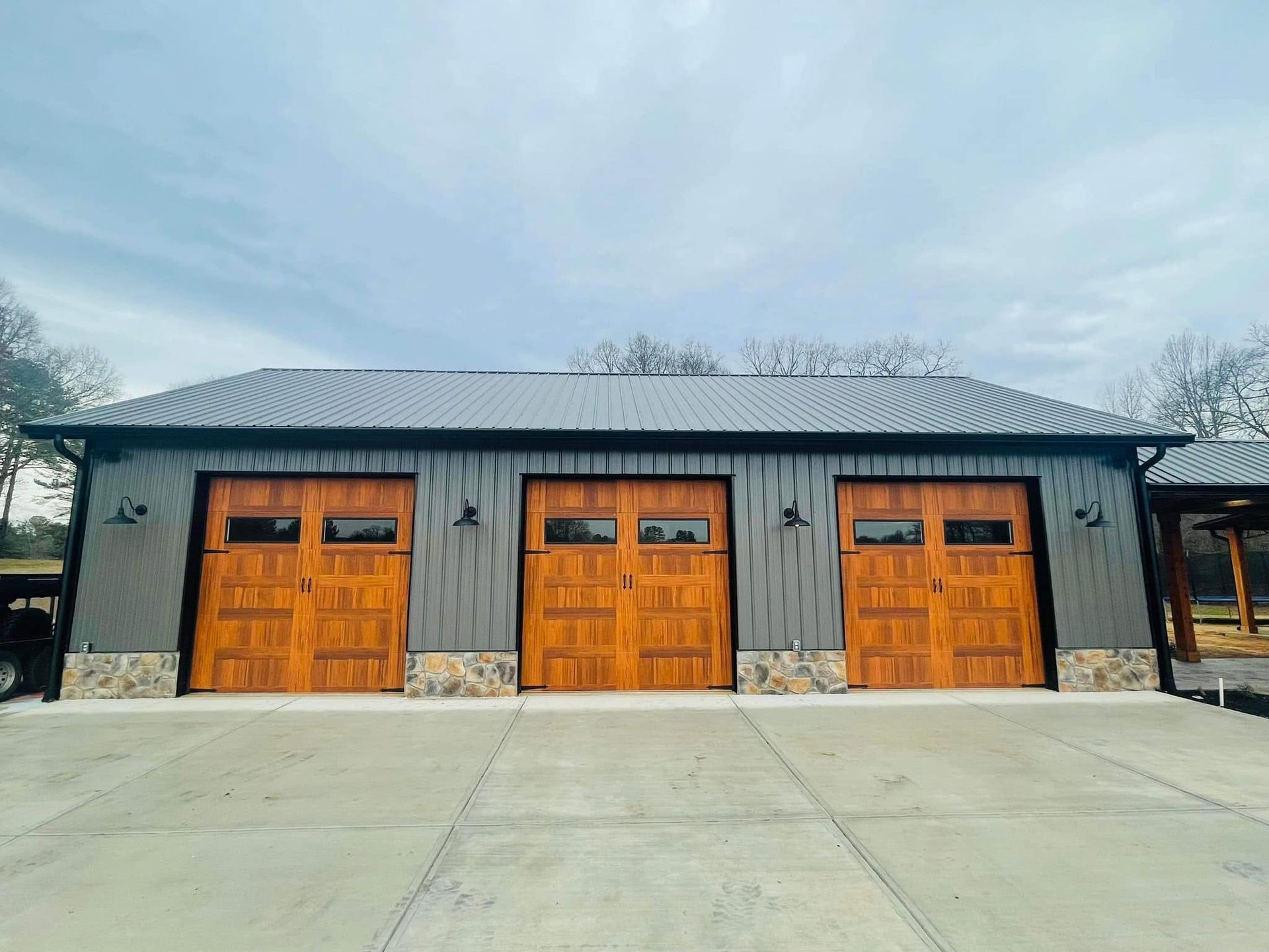 Three orange garage doors on a gray stone building with a dark metal roof and wide concrete driveway.