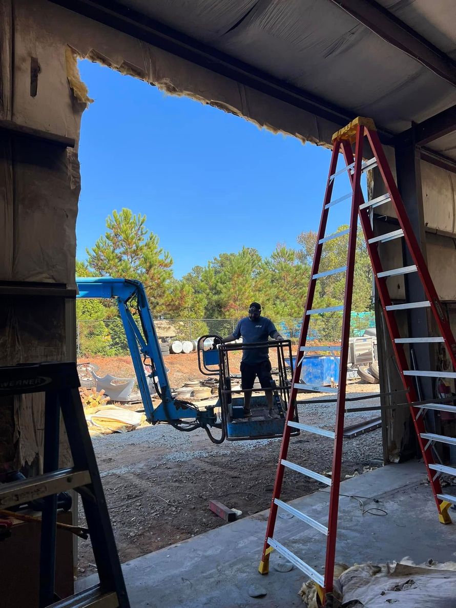 Construction site interior with a worker near ladders and wheelbarrow, seen through a large doorway.
