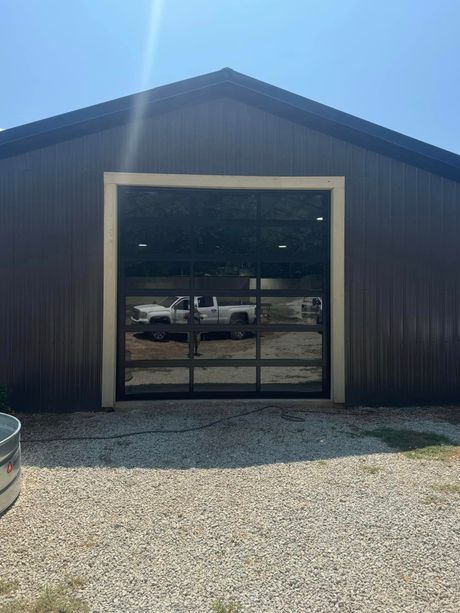 Dark metal building with a large glass garage door and gravel foreground under bright sun.