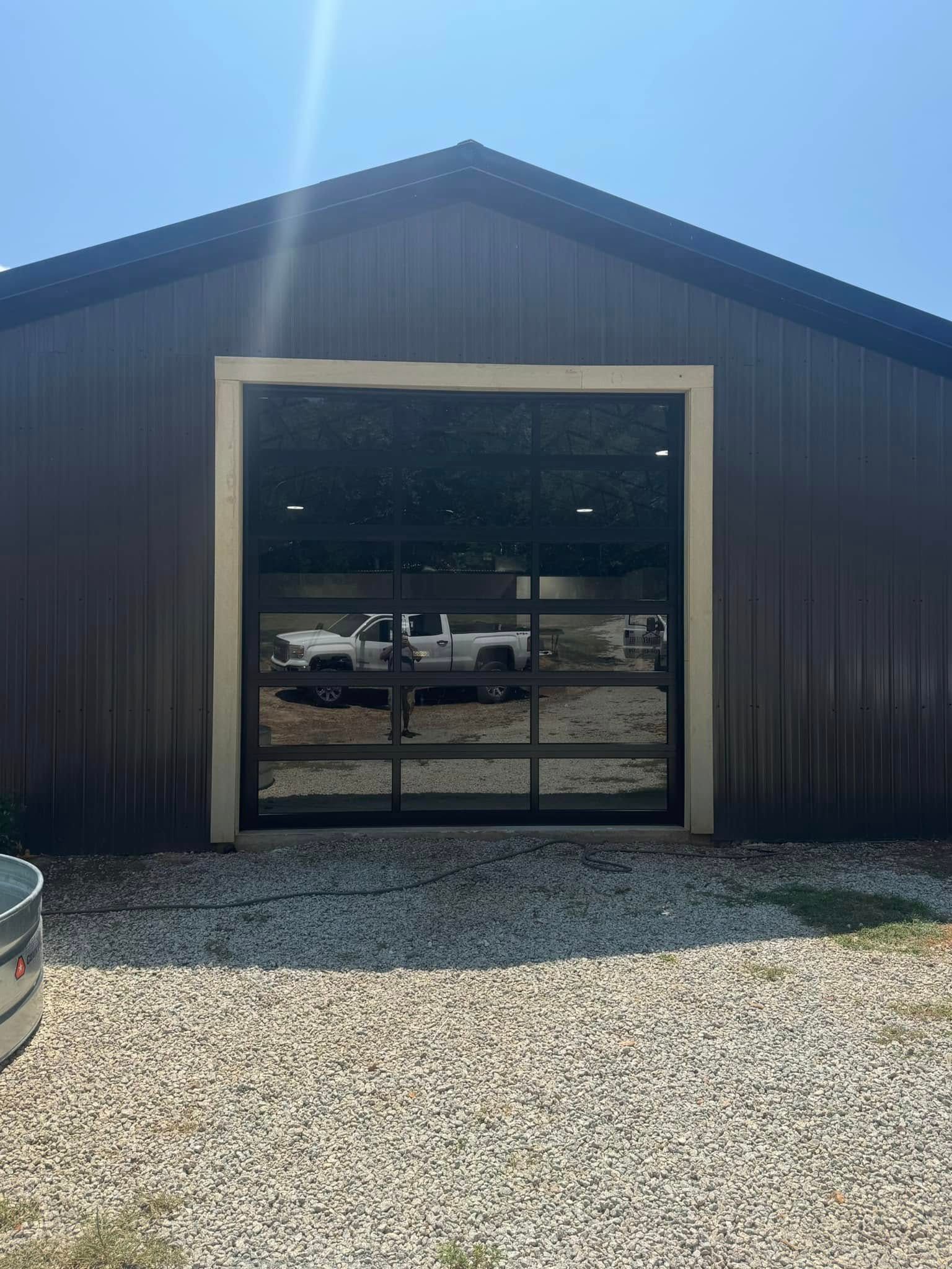 Dark metal building with a large glass garage door on a gravel lot in bright sunlight