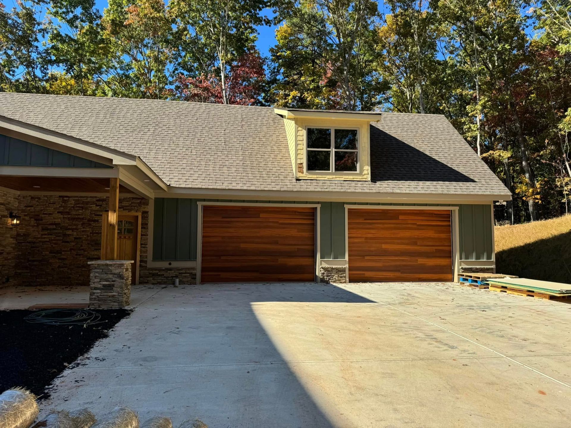 Suburban house with two wooden garage doors, gray roof, and sunlit driveway under trees