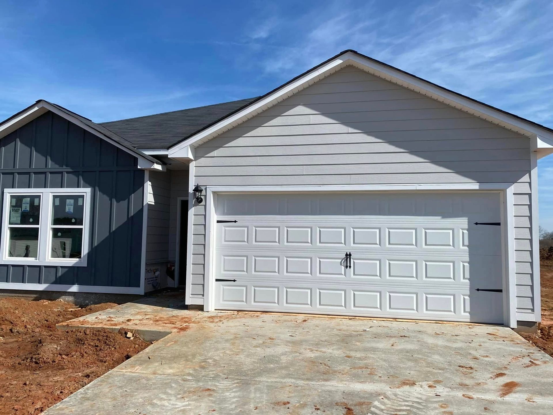 New single-story house with gray siding, white garage door, and unfinished driveway under a blue sky