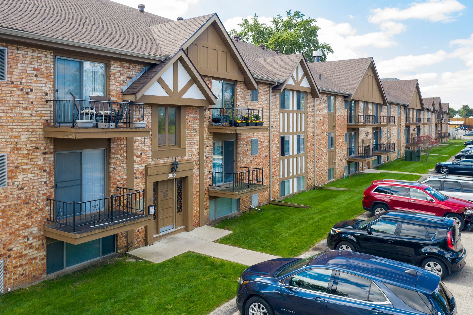 Apartment complex with brown brick, Tudor-style facade, and cars parked along the curb on a sunny day.