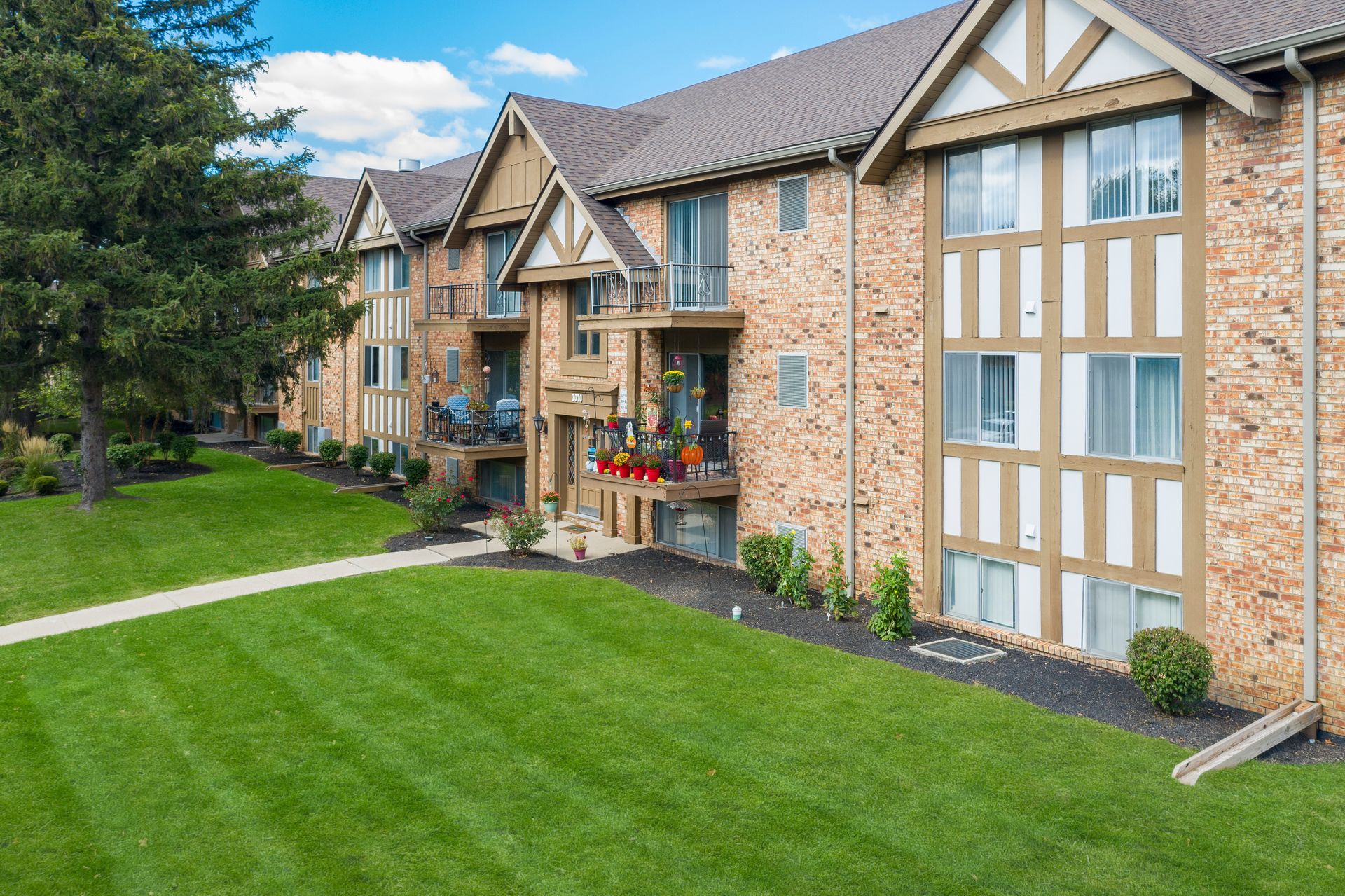 Apartment building with brick facade, brown roof, and green lawn. Balconies with people.