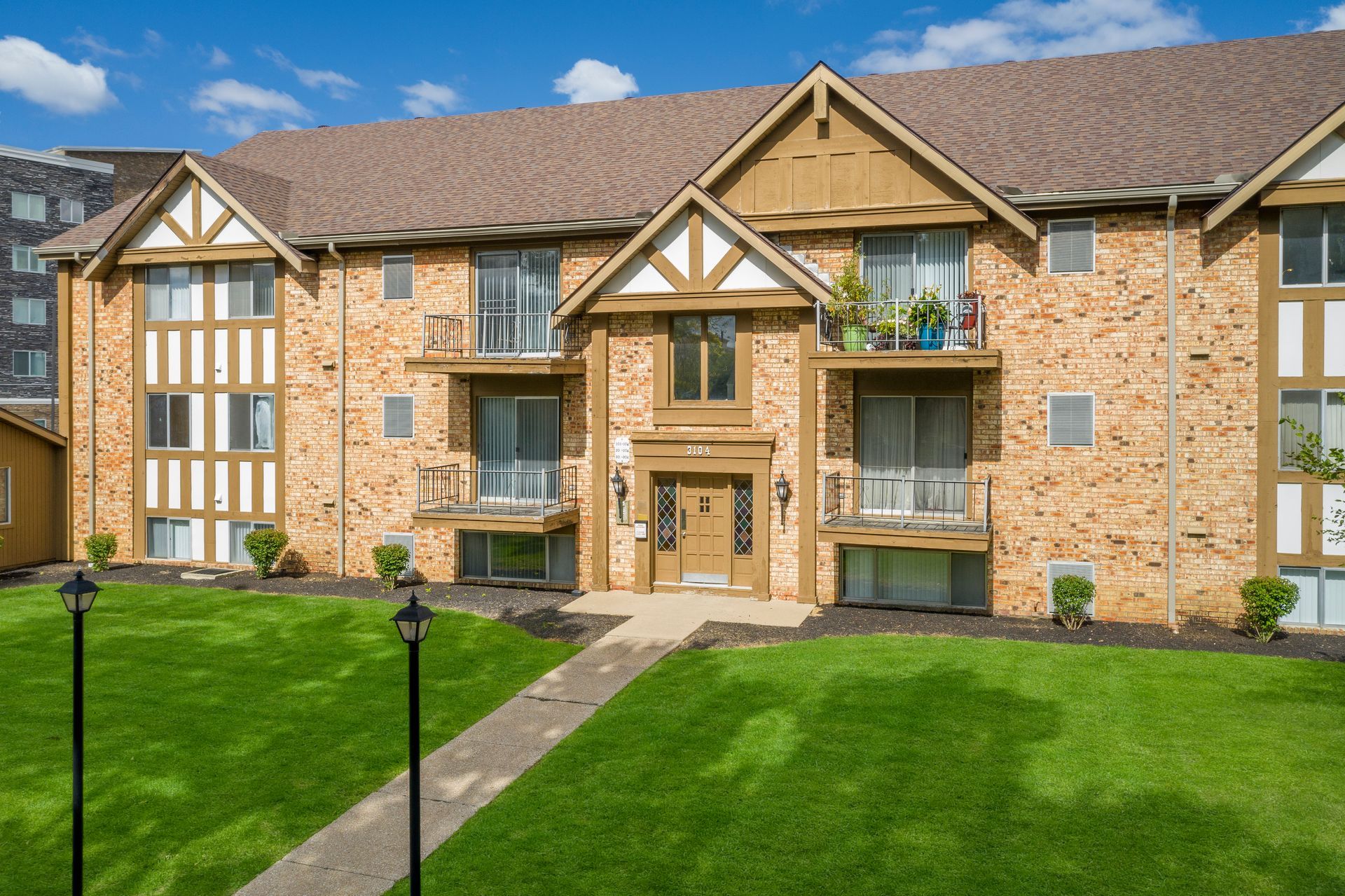 Apartment building with brown brick facade, gabled roof, balconies, and green lawn.