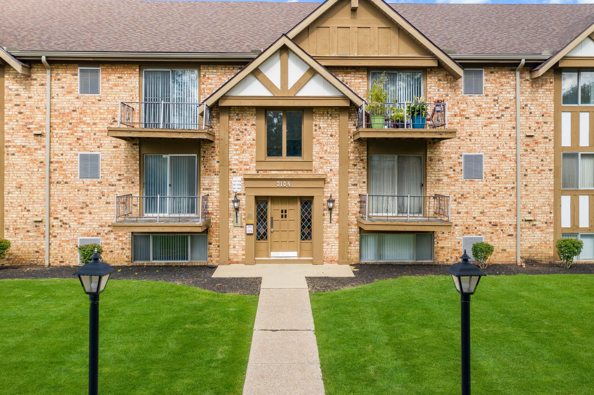 Apartment building exterior, brick and brown trim. Pathway leads to front door, two black lampposts on green lawn.