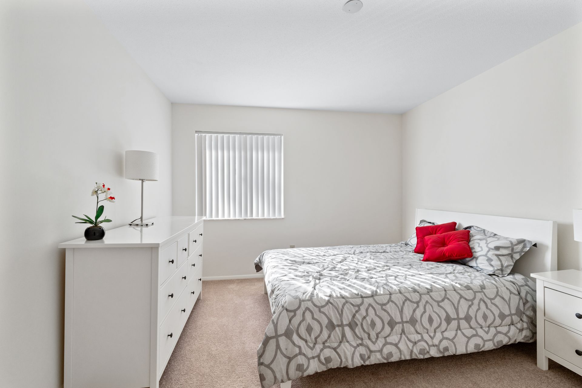 Bedroom with a white dresser and nightstand, queen-sized bed with gray patterned comforter, red pillows, and window with blinds.