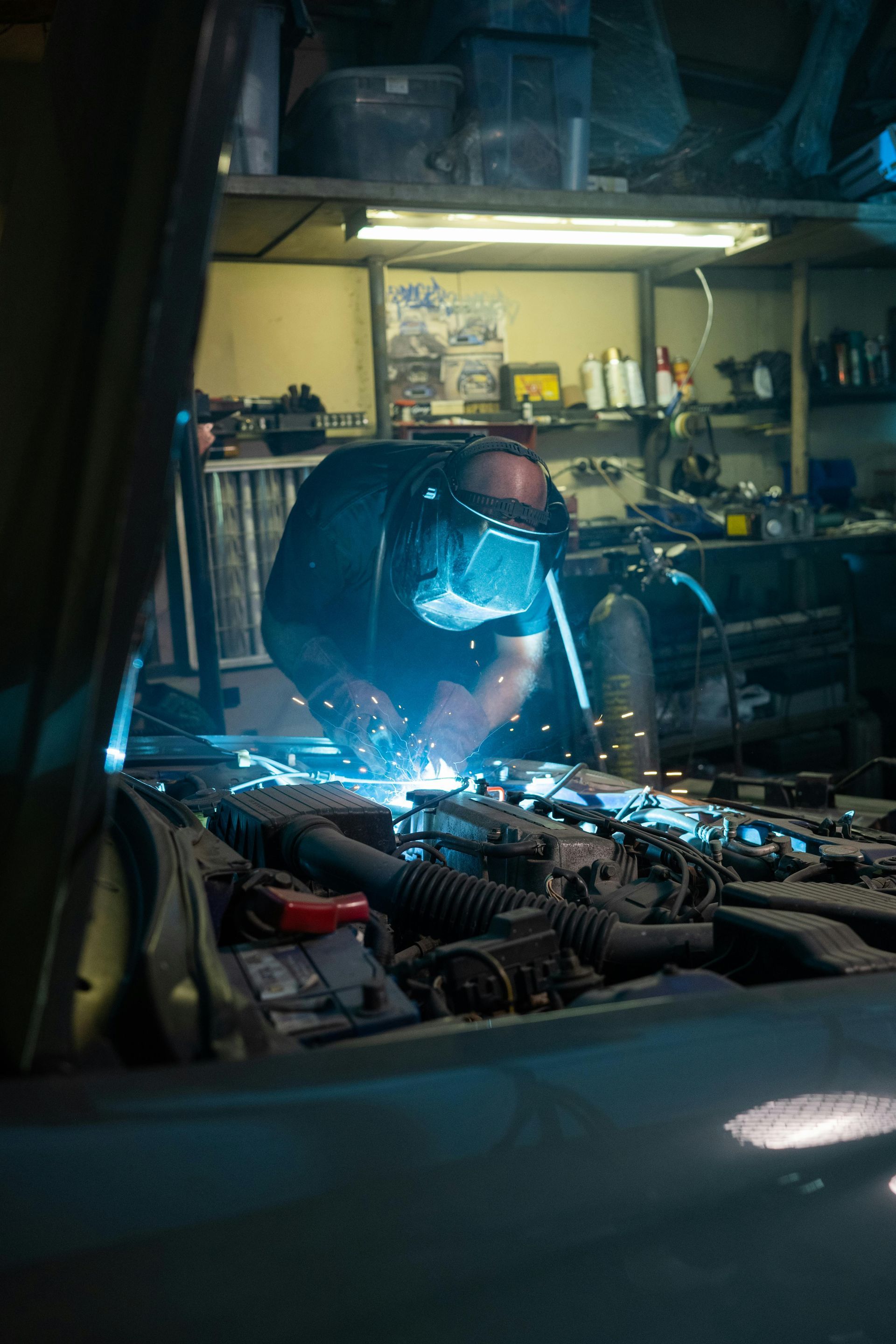 Man welding under the hood of a car in a cluttered garage, sparks flying. | KAP Motorsports