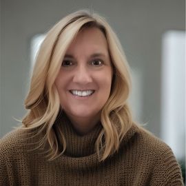 Woman with long blonde hair smiles, wearing a pink blazer and white shirt against a gray backdrop.