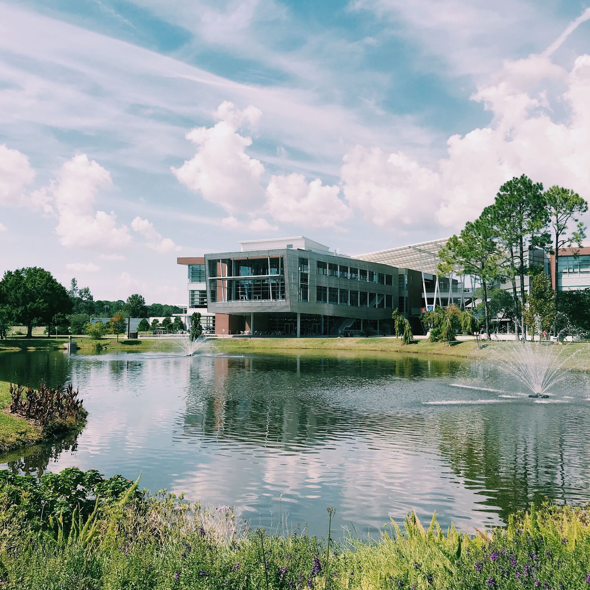 Modern office building beside a pond with fountains, framed by trees and a bright cloudy sky