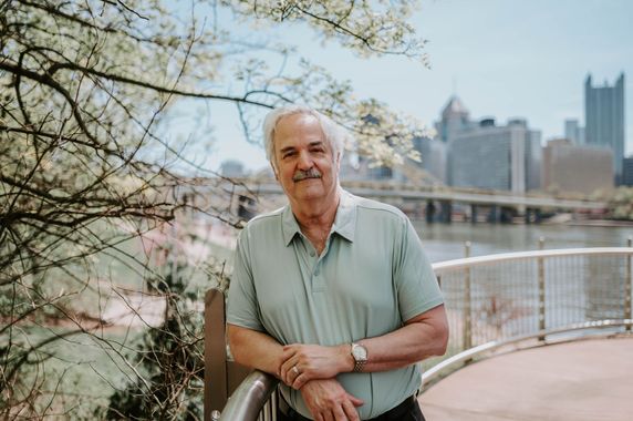 Man leaning on a railing in a park, with city buildings and pink blossoms in the background.