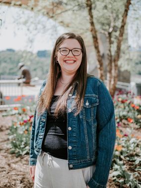 Smiling woman in glasses and denim jacket standing outdoors in a garden-like setting