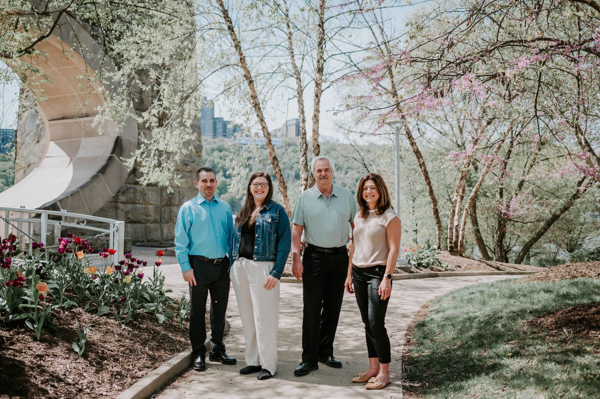 Four people standing on a garden path among blooming trees and flowers, smiling outdoors