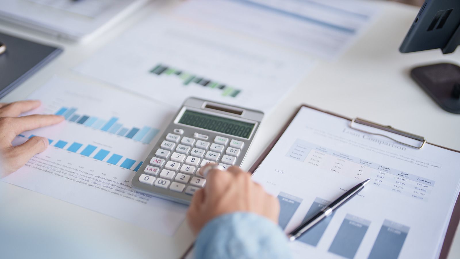 Hand calculating finances with a calculator beside charts and documents on a desk