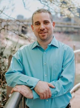 Man in a light blue shirt leaning on a railing outdoors near a body of water