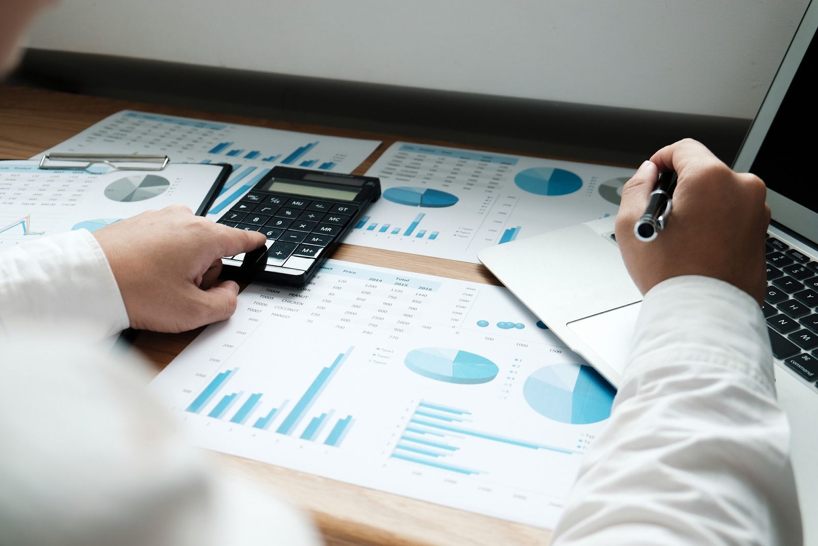 Two people reviewing blue charts and a calculator on a desk, one taking notes with a pen.