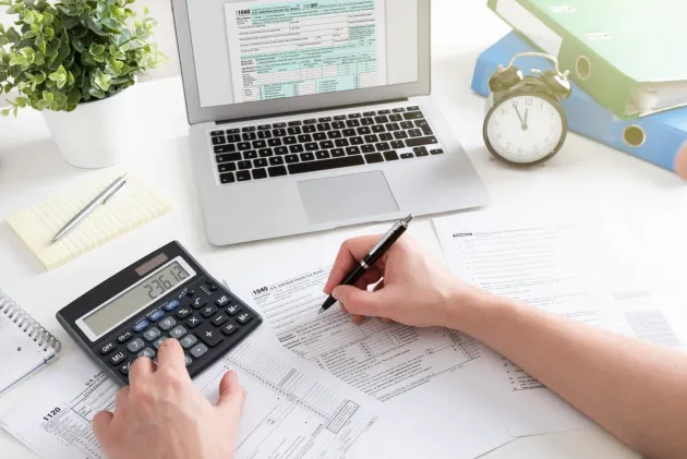 Hands calculating bills beside a laptop, calculator, and paperwork on a bright desk