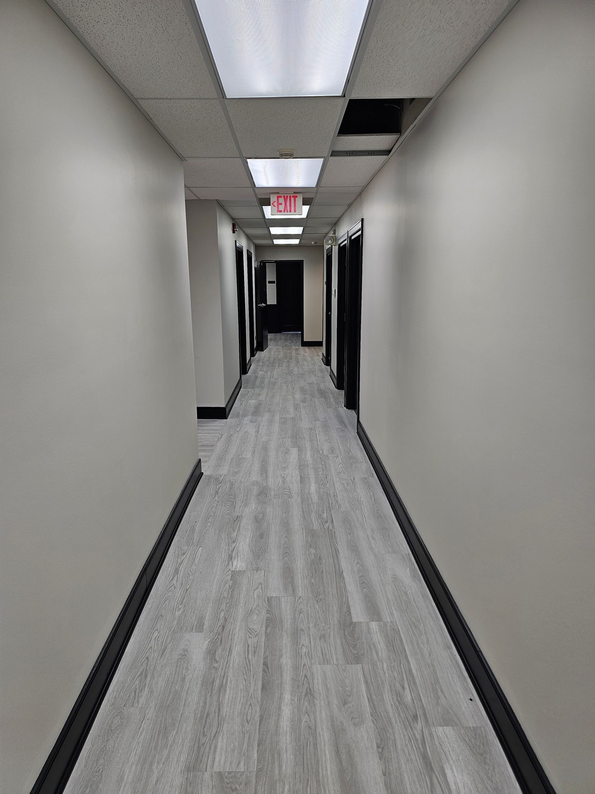 Long, narrow office hallway with gray walls, wood-look flooring, and overhead fluorescent lights.