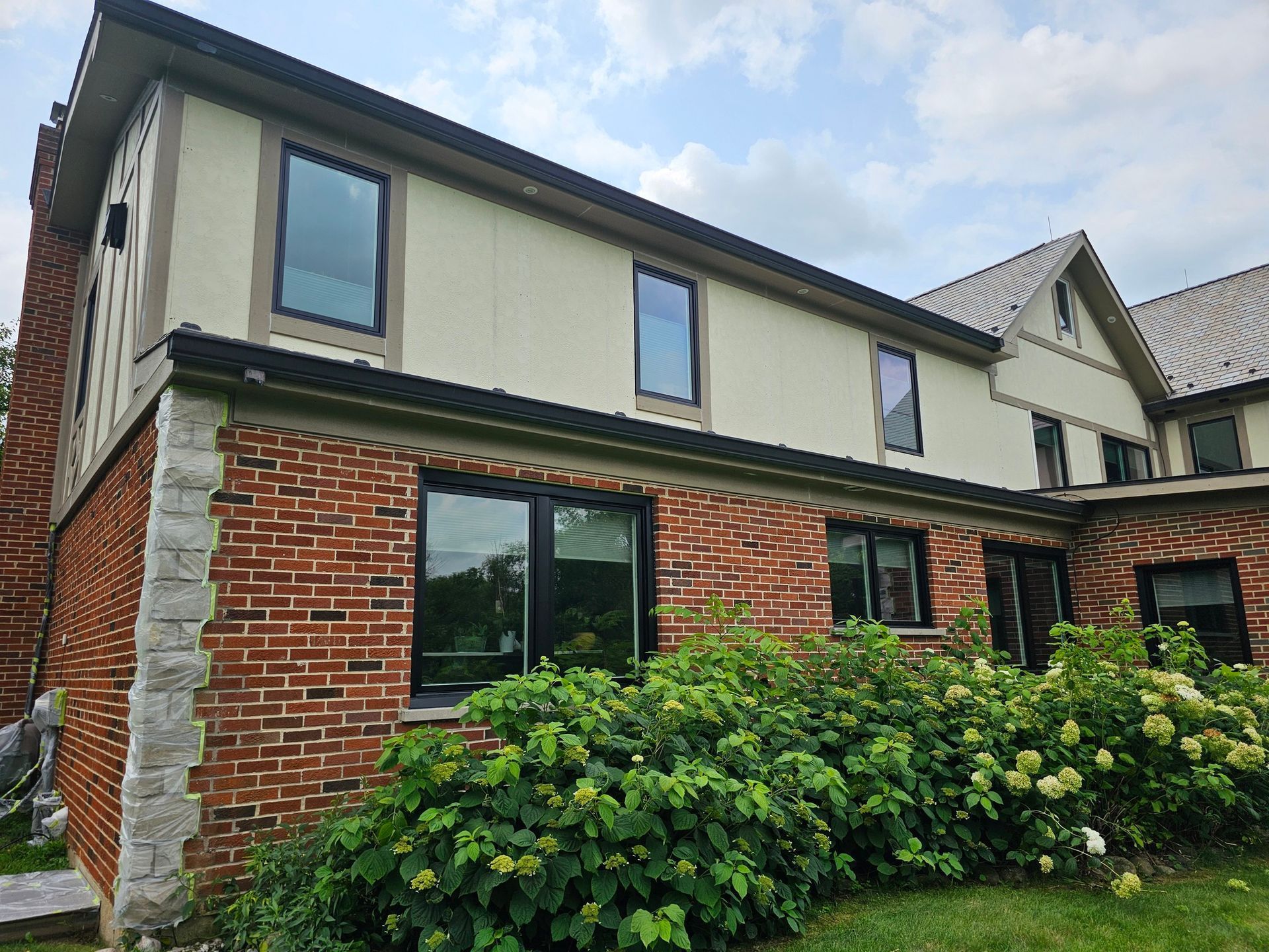 Two-story house with brick facade, cream siding, dark windows, and green shrubbery in front.