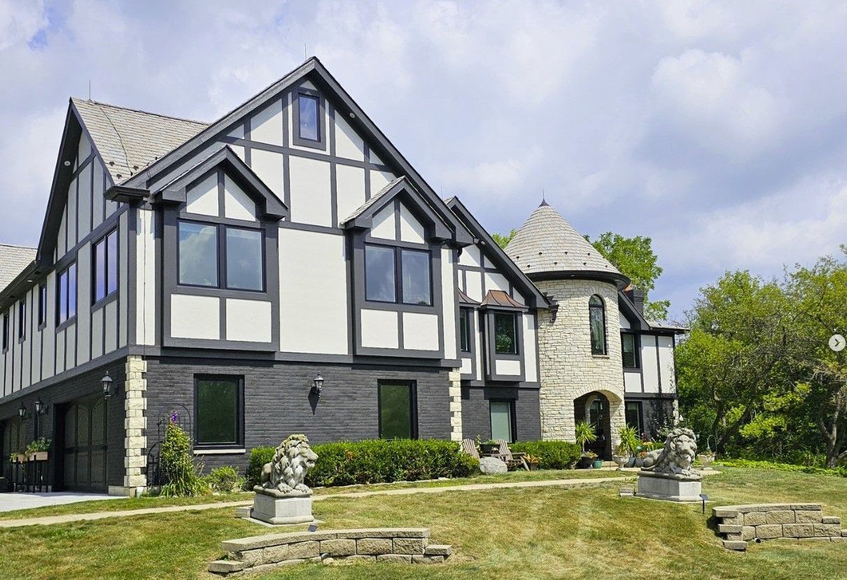 Tudor-style home with black and white facade, stone tower, and manicured lawn under a cloudy sky.