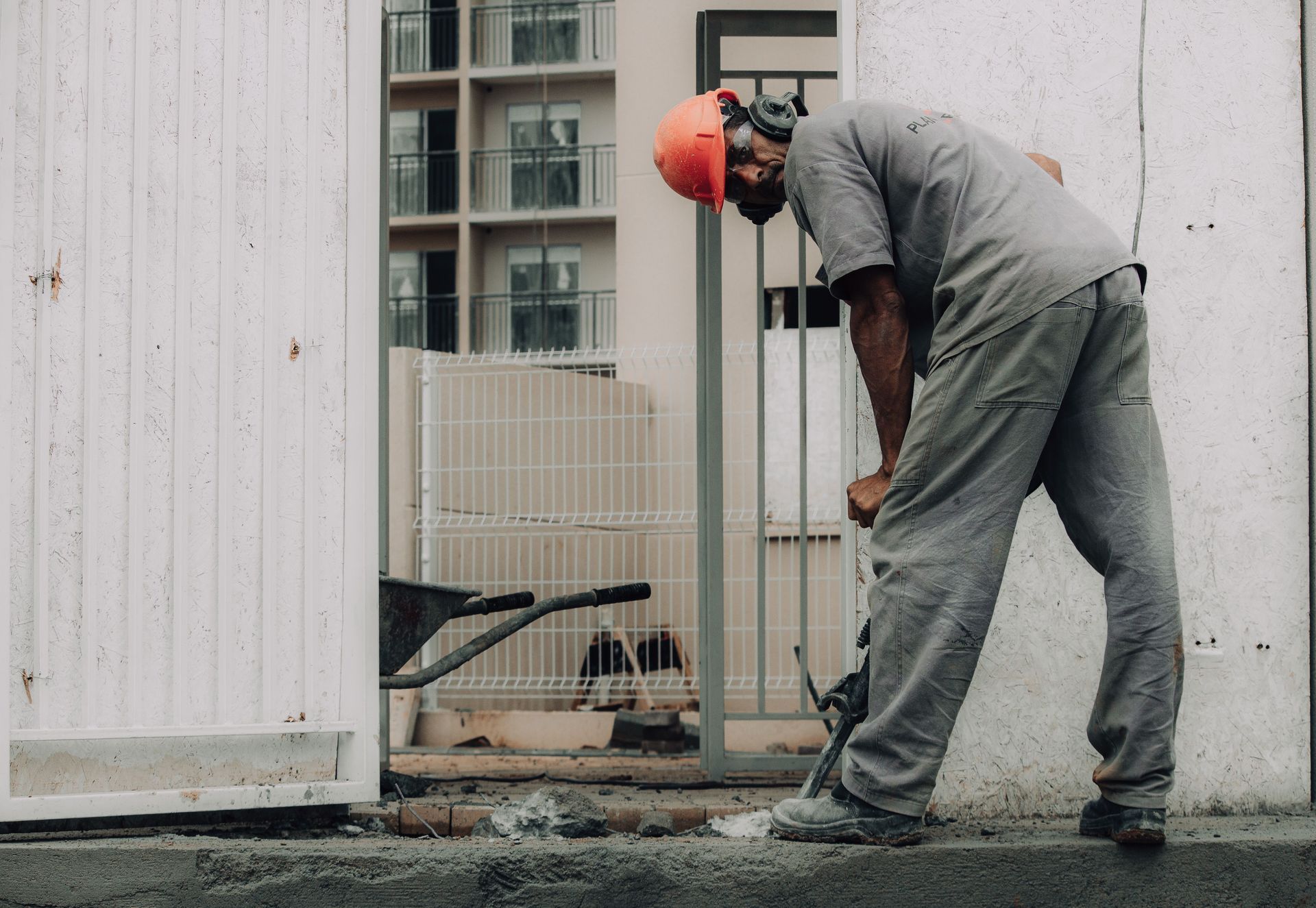 Construction worker in gray uniform and orange hard hat leaning forward beside a building wall.