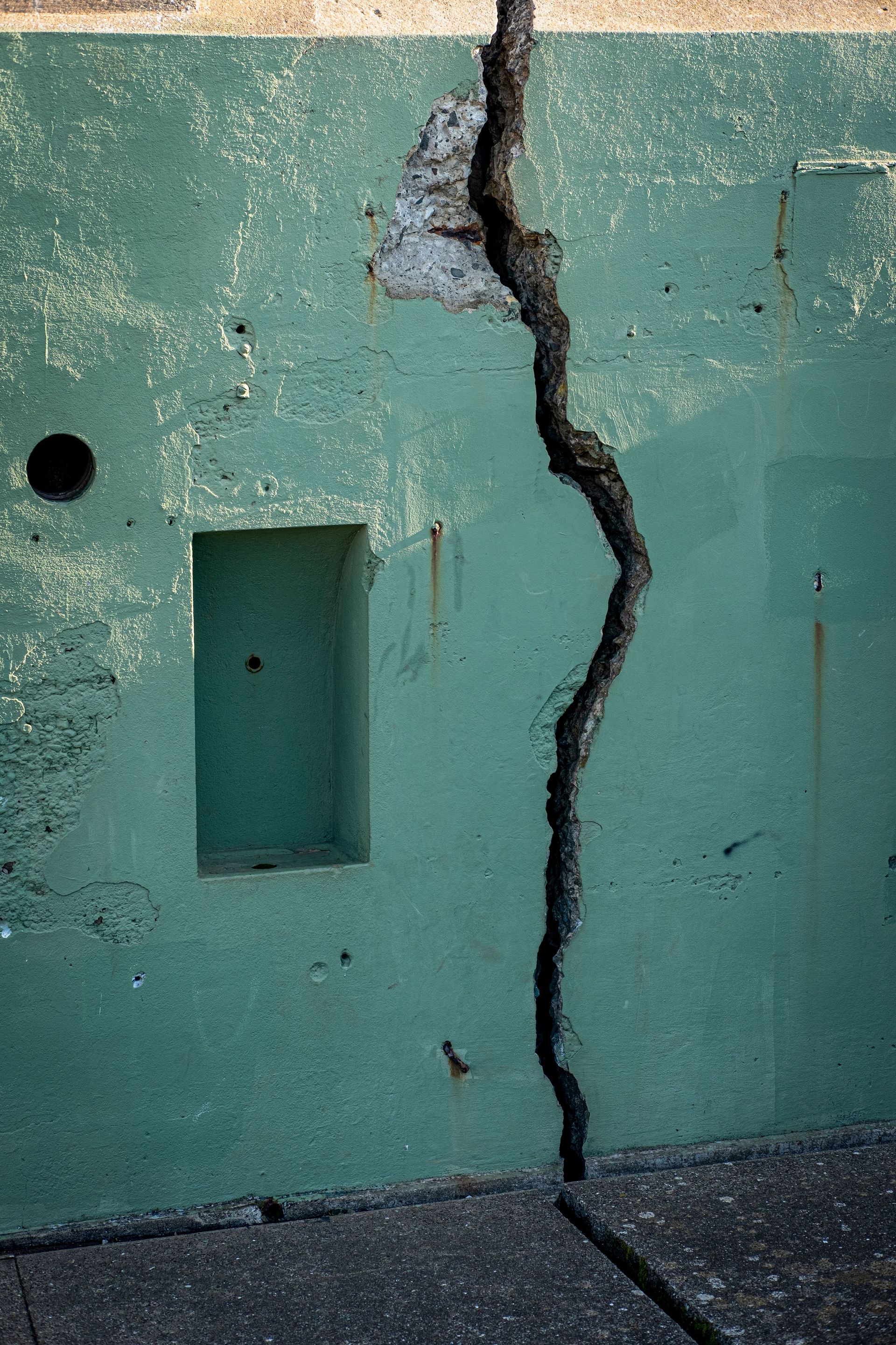 Cracked turquoise wall with a large vertical fissure and damaged concrete near the top