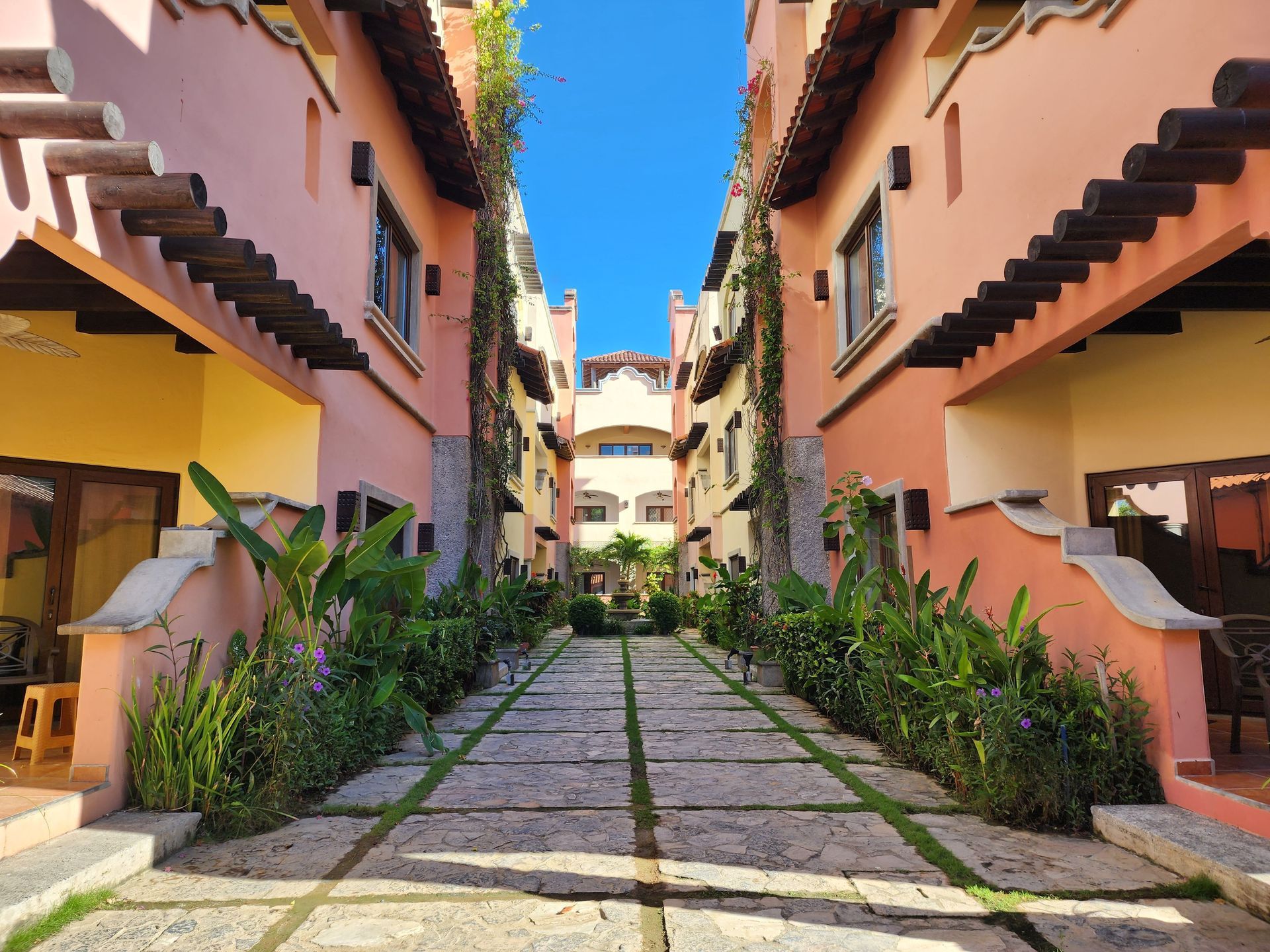 Symmetrical courtyard walkway between peach buildings with plants and a blue sky overhead