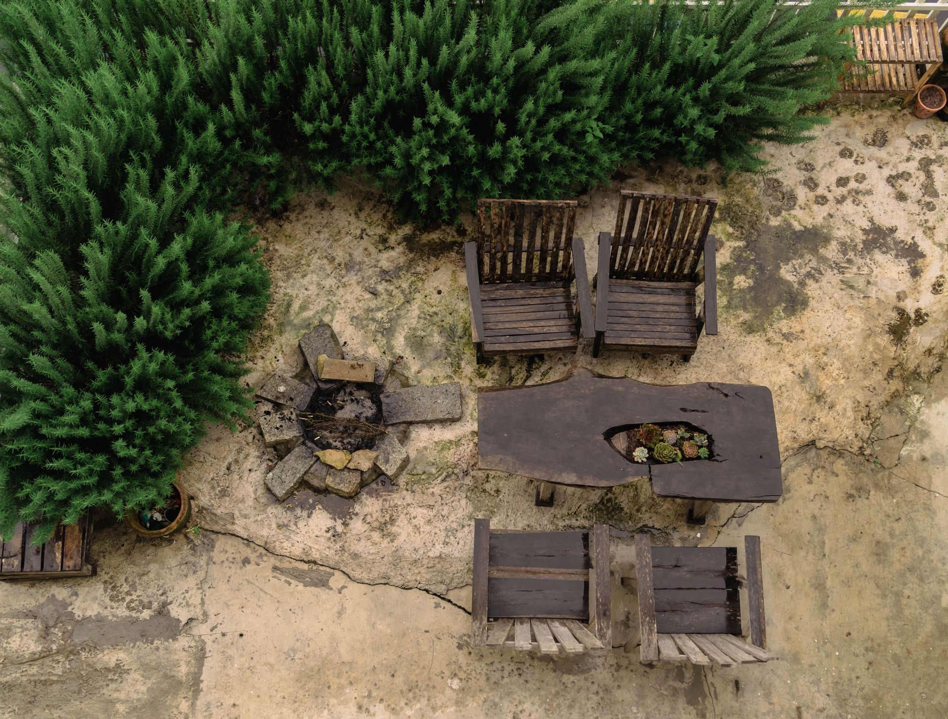 Aerial view of a patio with two chairs, a table, benches, and a fire pit beside green shrubs.