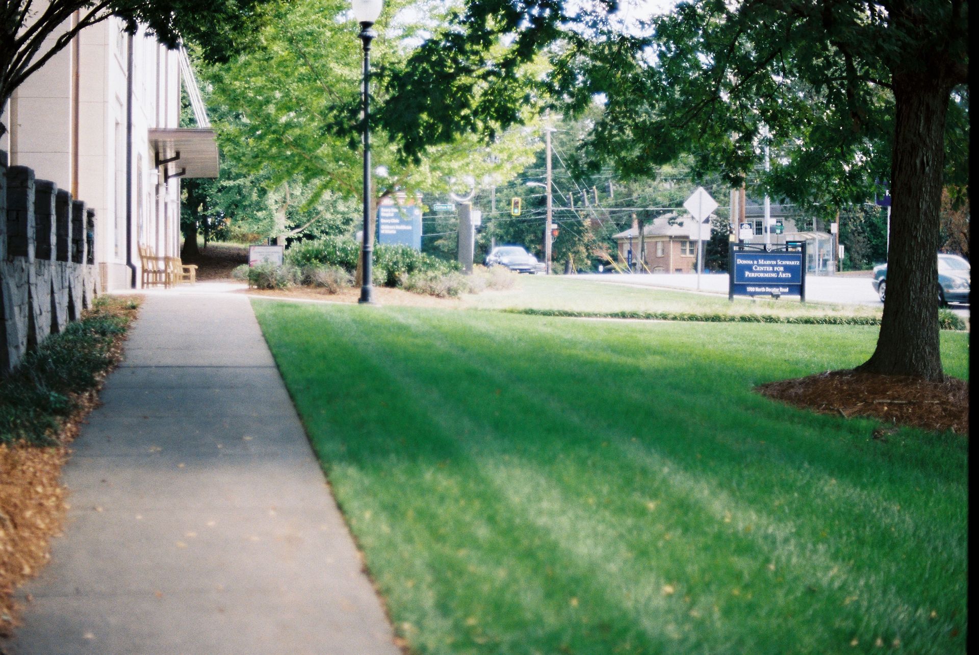 Sidewalk beside a building and green lawn with trees and a distant blue sign on a sunny day